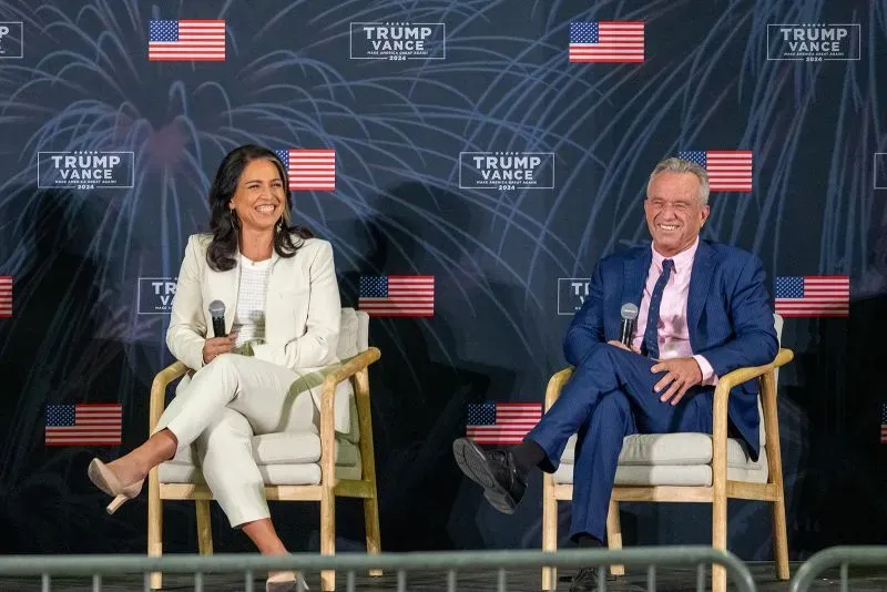 تظهر الصورة تولسي غابارد وروبرت كينيدي جونيور يجلسان معًا في حدث سياسي، مع خلفية تحمل شعار "ترامب فانس 2024" وأعلام أمريكية.