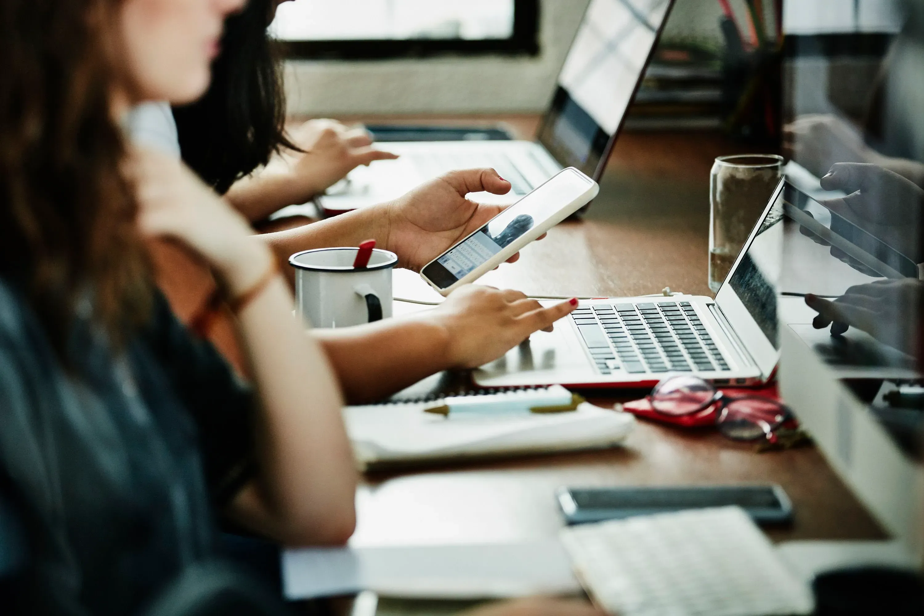 أشخاص يعملون على أجهزة الكمبيوتر المحمولة في مكتب، مع شخص يستخدم الهاتف الذكي، مما يعكس استخدام الذكاء الاصطناعي في بيئة العمل.