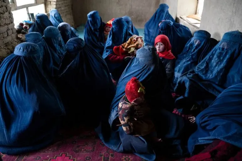 نساء يرتدين البرقع الأزرق يجلسن مع أطفالهن في غرفة، يعكس الصورة معاناة النساء والفتيات تحت حكم طالبان.