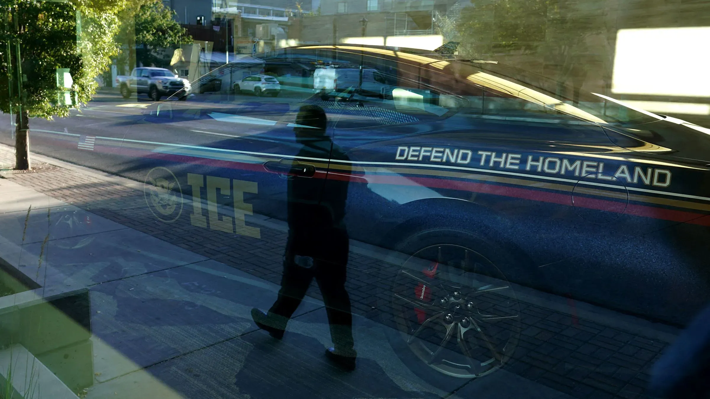 صورة تعكس ضابطًا يمشي بجانب سيارة تابعة لوكالة إنفاذ قوانين الهجرة والجمارك، مكتوب عليها "دافع عن الوطن".