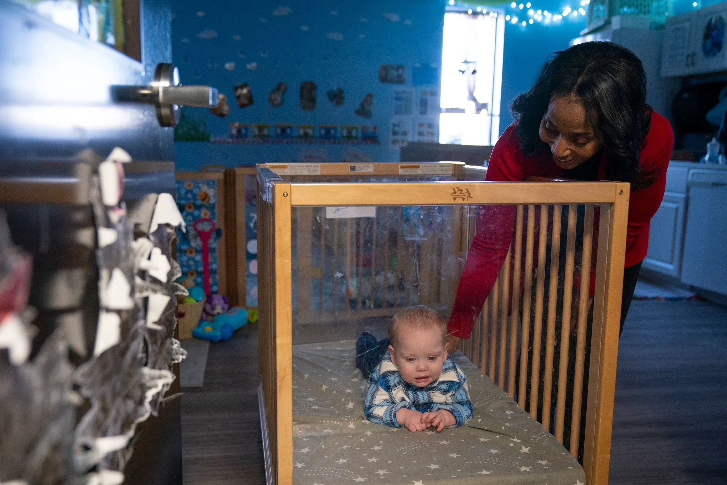 امرأة تراقب طفلاً صغيراً داخل سرير أطفال زجاجي في مركز رعاية نهارية، مع خلفية ملونة وألعاب، تعكس أهمية رعاية الأطفال.