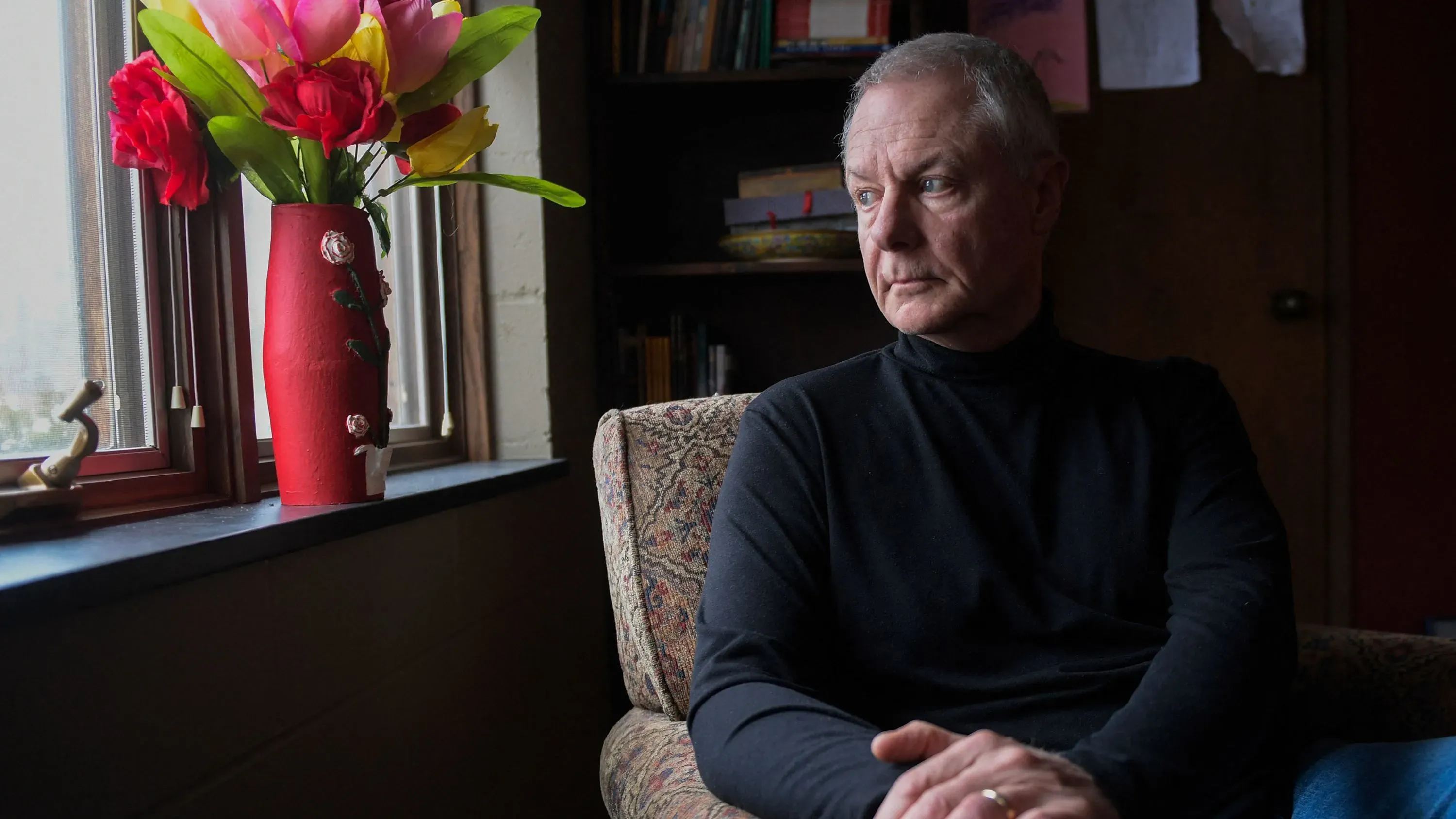 رجل مسن يجلس بجوار نافذة مضاءة، يتأمل بجدية، مع زهور ملونة في مزهرية حمراء بجانبه، مما يعكس أجواء من التأمل والقلق.