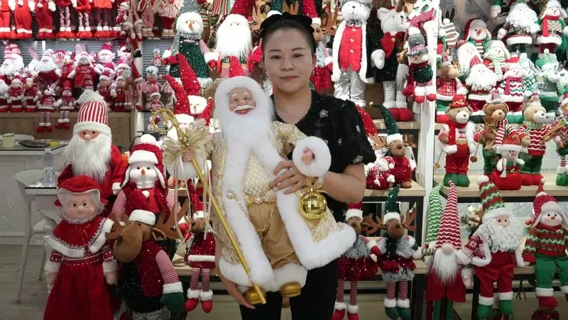 امرأة تحمل دمية سانتا في متجر مليء بزينة عيد الميلاد، تعكس تأثير مدينة ييوو الصينية على سوق الزينة في الولايات المتحدة.