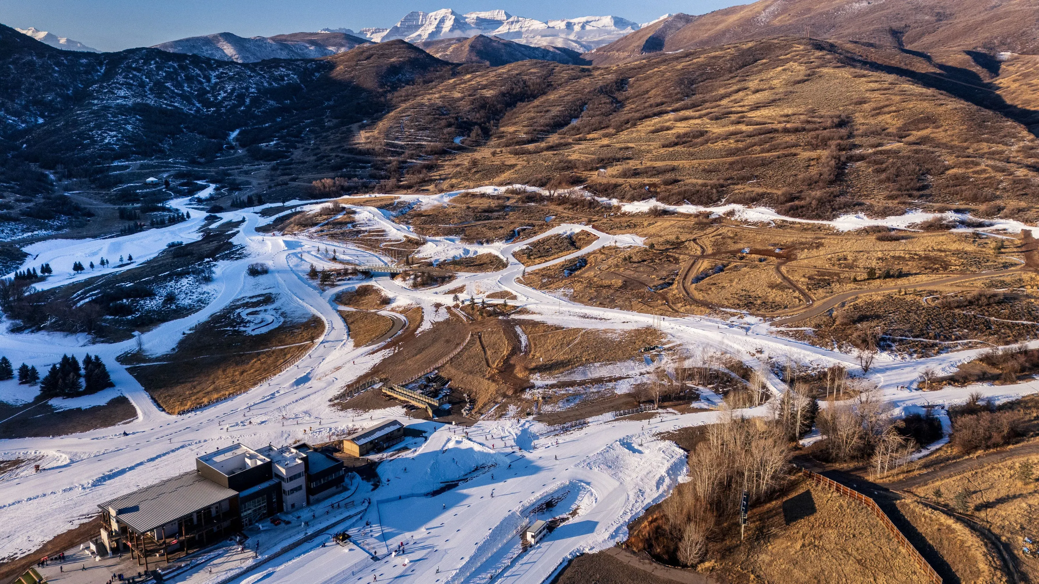 منظر جوي لموقع رياضي في جبال الألب الإيطالية، يظهر مسارات التزلج مع ثلوج صناعية وأرض جافة، مما يعكس تأثير تغير المناخ على الرياضات الشتوية.