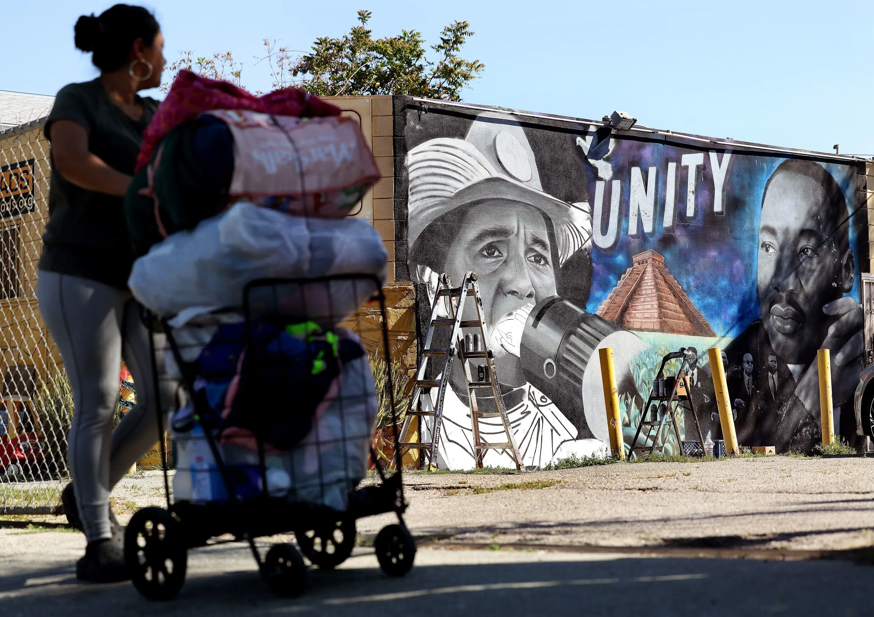 امرأة تدفع عربة مليئة بالأشياء بالقرب من جدارية تصور شخصيات تاريخية بارزة، مع التركيز على الوحدة والتضامن.