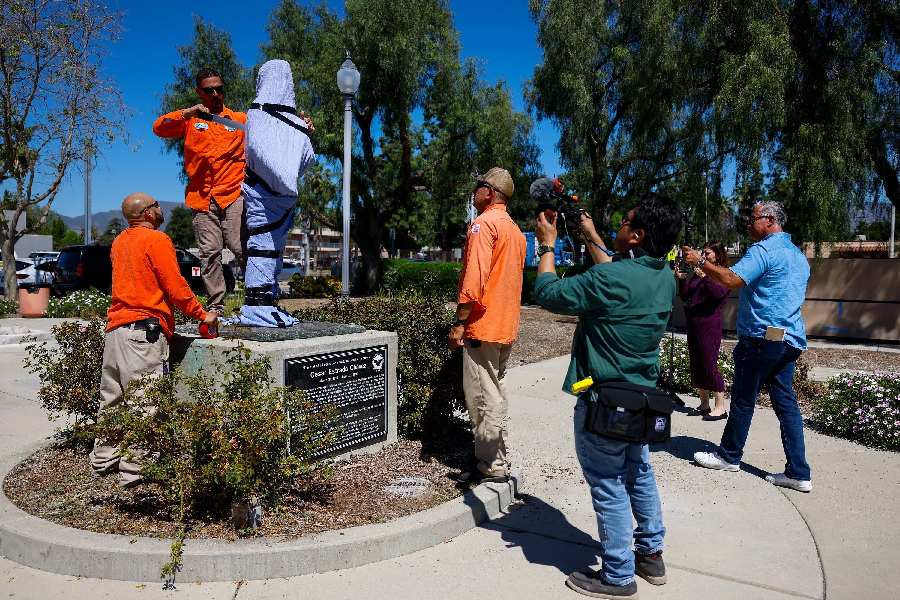 عمال يقومون بتغطية تمثال سيزار تشافيز في حديقة، بينما يلتقط الصحفيون الصور. يعكس الحدث تراجع إرثه كرمز للحقوق المدنية.