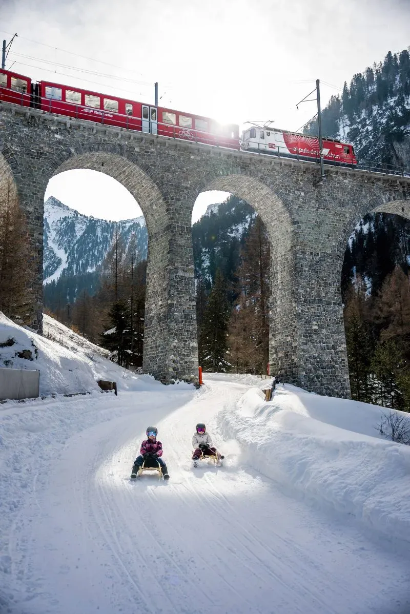 طفلان يتزلجان على زلاجات خشبية في مسار ثلجي، مع قطار أحمر يمر فوق جسر حجري في جبال الألب السويسرية.