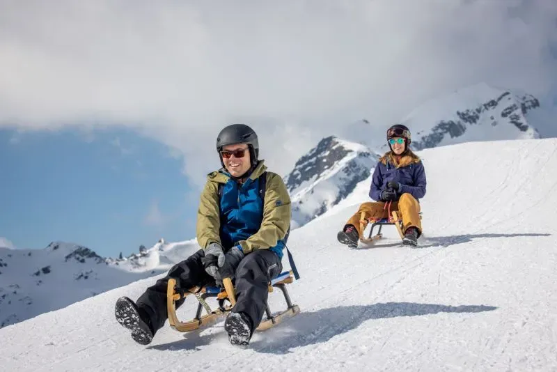 شخصان يرتديان خوذات يتزلجان على زلاجات خشبية في جبال الألب السويسرية، مع مناظر جبلية خلابة في الخلفية.