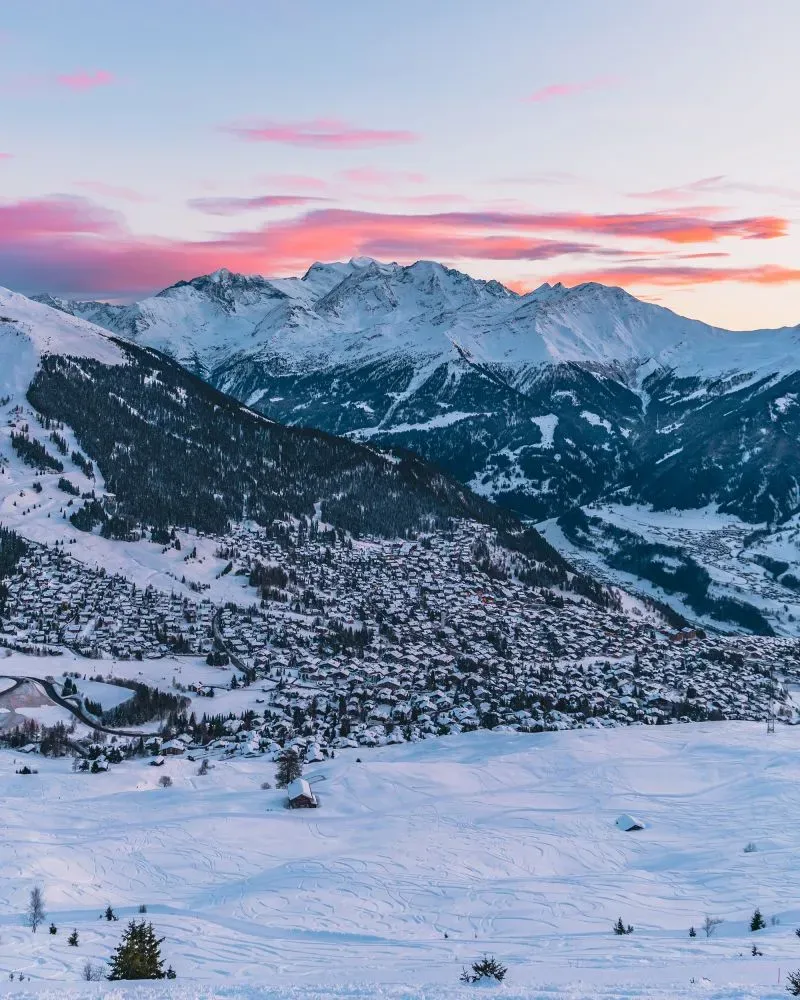 منظر جوي لقرية جريندلوالد السويسرية محاطة بالجبال المغطاة بالثلوج، مع غروب الشمس الذي يضيء السماء بألوان دافئة.