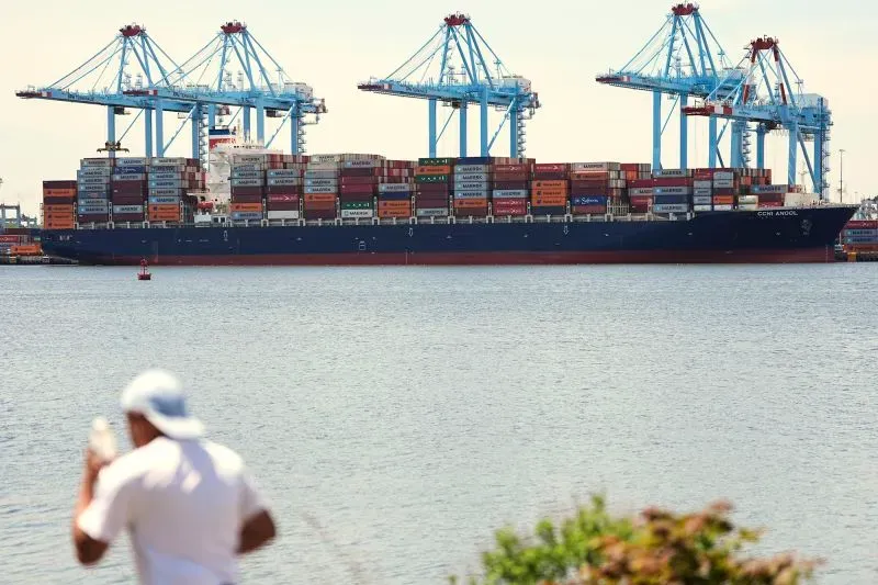 سفينة شحن محملة بالحاويات في ميناء، مع وجود رافعات خلفية، تعكس تأثير التجارة الدولية والتعريفات الجمركية على الاقتصاد.