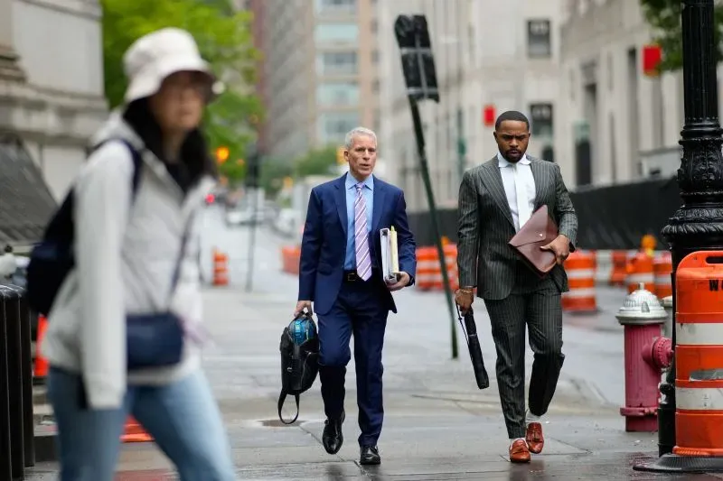 محاميان يتجهان نحو المحكمة في مدينة نيويورك، يحمل أحدهما حقيبة وثائق والآخر مظلة، مع وجود عوائق مرورية في الخلفية.