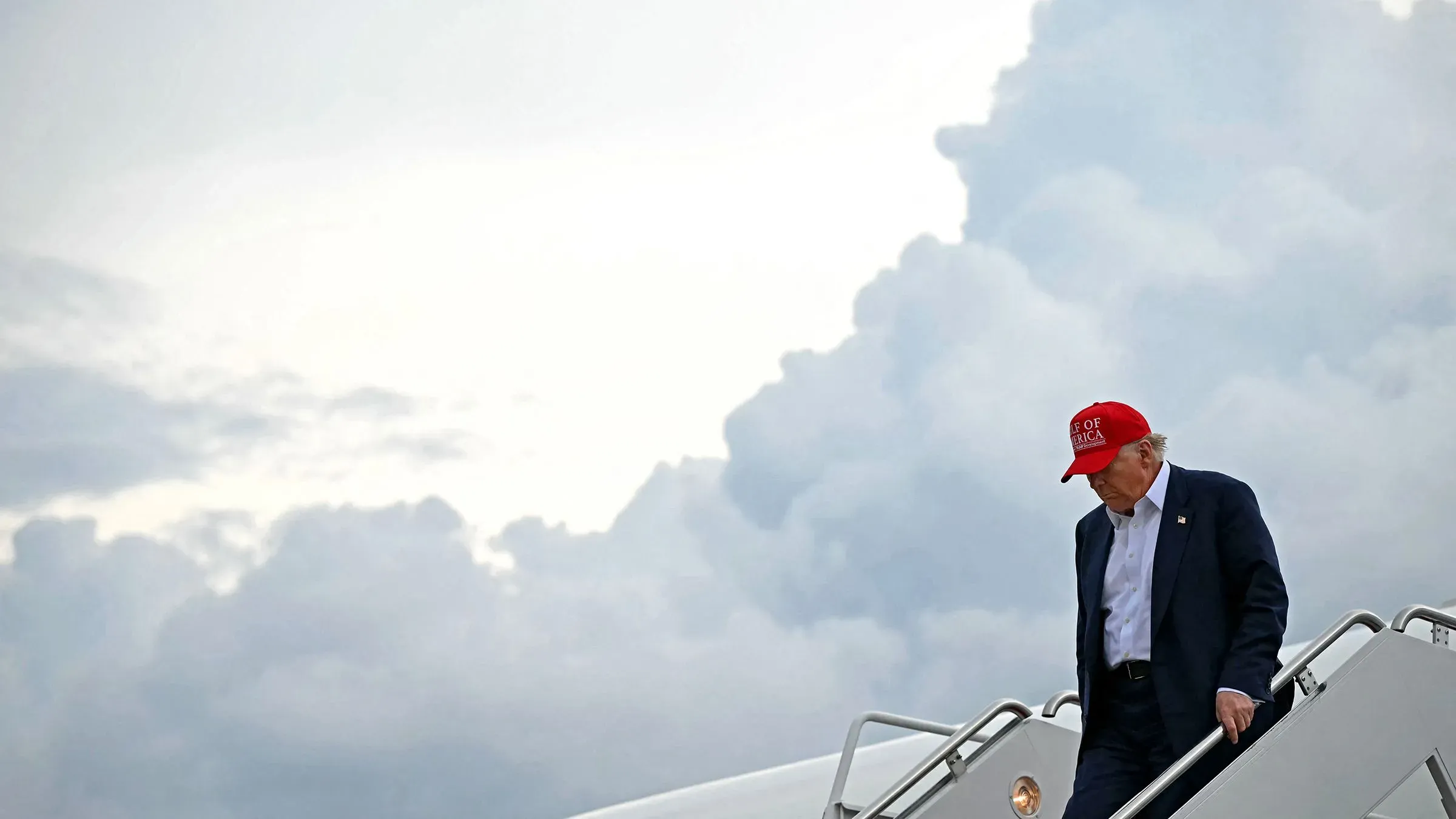 ترامب ينزل من الطائرة الرئاسية مرتديًا قبعة حمراء، مع سماء غائمة في الخلفية، بعد ساعات من تمرير مشروع قانون مهم.