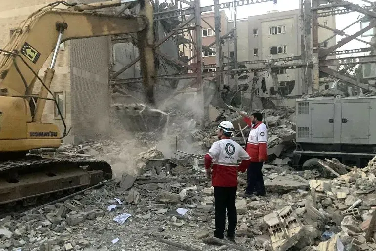 عمال إنقاذ يتفقدون موقعًا مدمرًا في طهران بعد غارة إسرائيلية، مع وجود جرافة وآثار دمار واسع في المباني المحيطة.