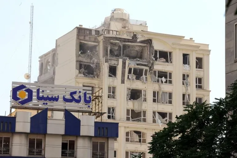 تظهر الصورة مبنى متضرر بشدة جراء الهجمات الإسرائيلية، مع لافتة لبنك سينا في المقدمة، مما يعكس الأثر المدمر على البنية التحتية الإيرانية.