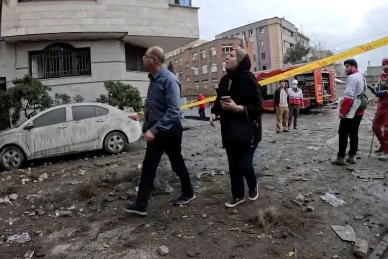 مشهد لعدد من الأشخاص يسيرون في منطقة متضررة في طهران، مع وجود سيارات إسعاف وخطوط تحذير، مما يعكس آثار الهجمات على البنية التحتية.