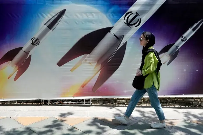 امرأة تسير بجانب لافتة تظهر صواريخ تحمل شعار إيران، تعكس التوترات العسكرية والتهديدات بين إيران والولايات المتحدة.