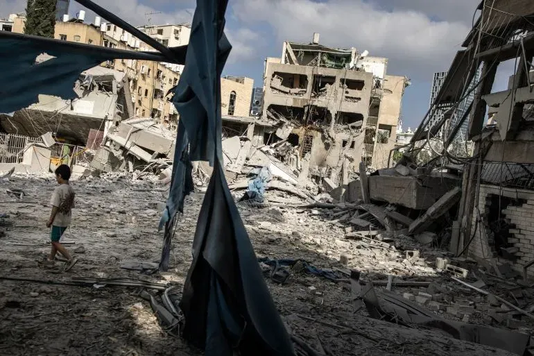 طفل يسير بين الأنقاض في منطقة تعرضت لقصف، مع مبانٍ مدمرة في الخلفية، مما يعكس آثار الصراع المستمر بين إسرائيل وإيران.