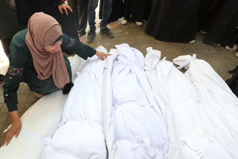 امرأة تبكي وتلمس أكفان ضحايا الغارات الإسرائيلية في غزة، بينما يتجمع الناس حولها في مشهد مؤلم يعكس حجم المأساة.