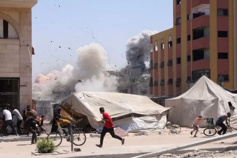 دخان يتصاعد من انفجارات في غزة، بينما يهرب الأطفال والبالغون من منطقة الهجوم، وسط خيام النازحين والمباني المدمرة.
