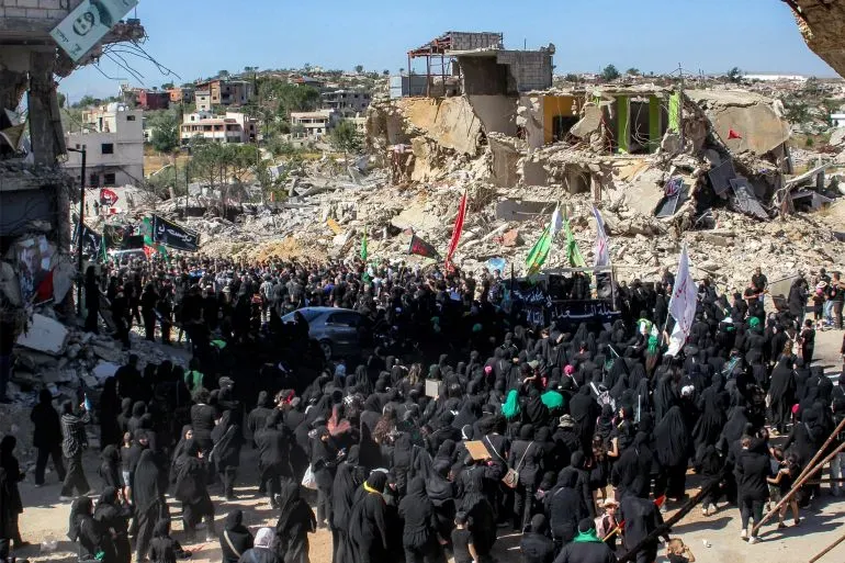 مظاهرة حاشدة في منطقة مدمرة، حيث تجمع المئات من الأشخاص بالزي الأسود، مع ظهور أعلام ملونة في الخلفية.