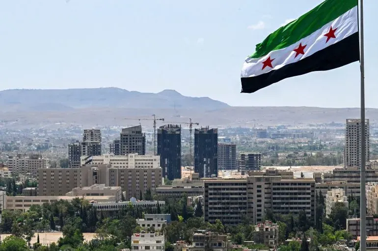 علم سوري يرفرف فوق مدينة دمشق، مع خلفية تظهر المباني الحديثة والجبال، تعكس الوضع المعقد في سوريا بعد النزاع المستمر.
