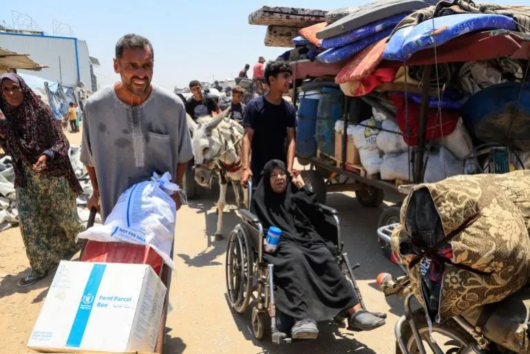 نازحون فلسطينيون يحملون الأمتعة ويعبرون منطقة حدودية، مع امرأة مسنّة على كرسي متحرك، في سياق أزمة إنسانية متفاقمة في غزة.