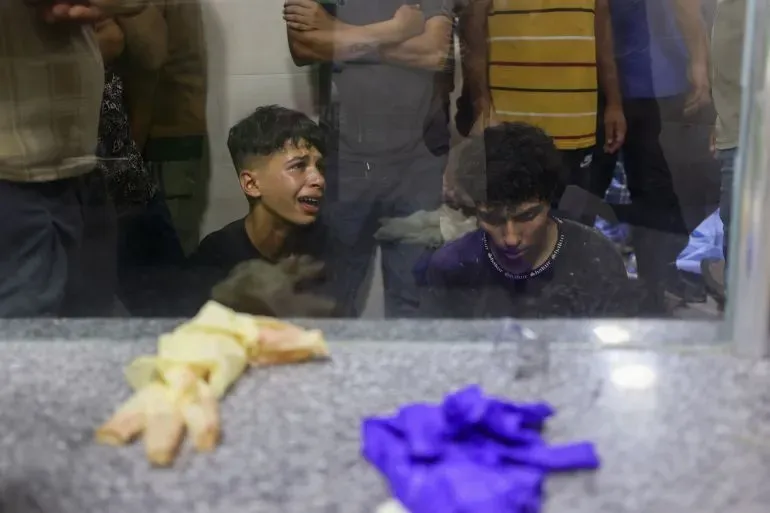 شبان يبكون أمام نافذة مستشفى، مع وجود أشخاص آخرين في الخلفية، بعد مقتل العديد من الفلسطينيين في هجمات إسرائيلية على مواقع توزيع المساعدات في غزة.