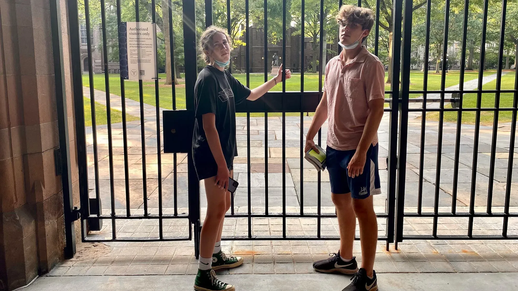 صورة لشابين يقفان أمام بوابة مغلقة في حرم جامعي، مع تواجد أشجار خضراء في الخلفية، تعكس شعور الانفصال والتباعد الاجتماعي.