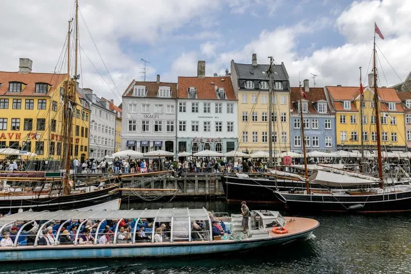 منظر لواجهة مائية في كوبنهاغن، يظهر مباني ملونة وسفن، مع تجمعات للناس في المطاعم والمقاهي، يعكس تأثيرات السفر في ظل التوترات السياسية.