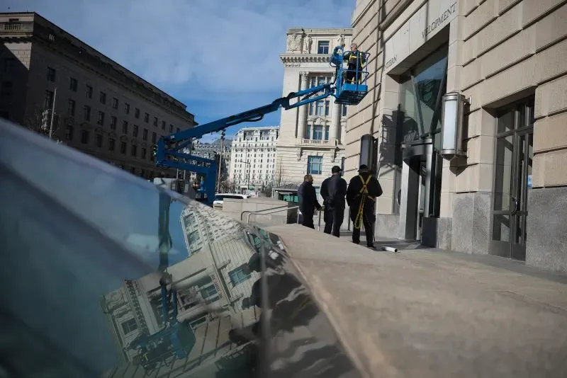 عمال يستخدمون رافعة لرفع المعدات في واجهة مبنى حكومي، مع ظهور أشخاص يقفون في الجهة المقابلة. الصورة تعكس التغيرات في الإدارة الحكومية.