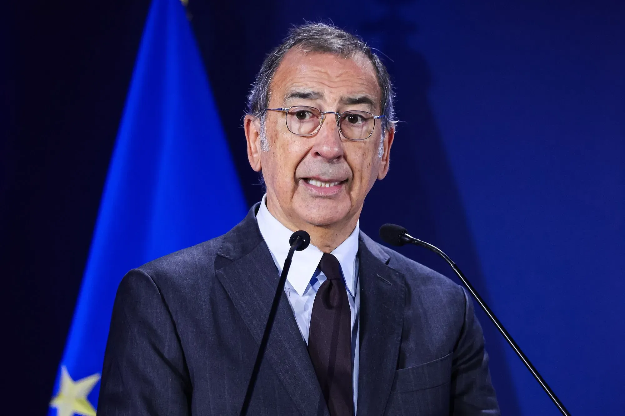 رجل يتحدث في مؤتمر صحفي مع العلم الإيطالي خلفه، يناقش وجود عملاء إدارة الهجرة والجمارك الأمريكية في أولمبياد ميلانو.