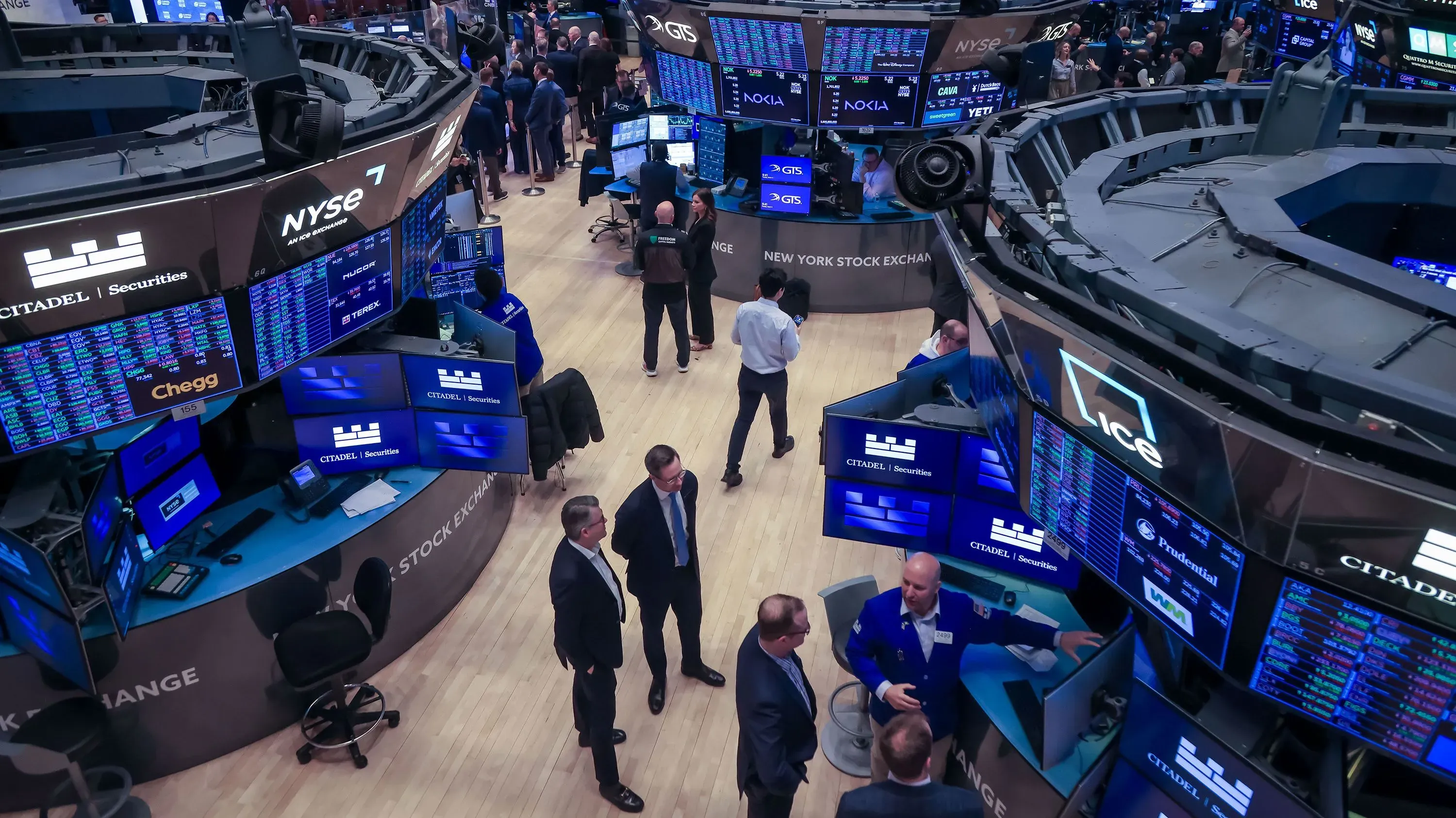 تظهر الصورة مشهدًا من بورصة نيويورك، حيث يتجمع المتداولون أمام شاشات تعرض أسعار الأسهم، مما يعكس حالة القلق في الأسواق بسبب سياسات ترامب الاقتصادية.