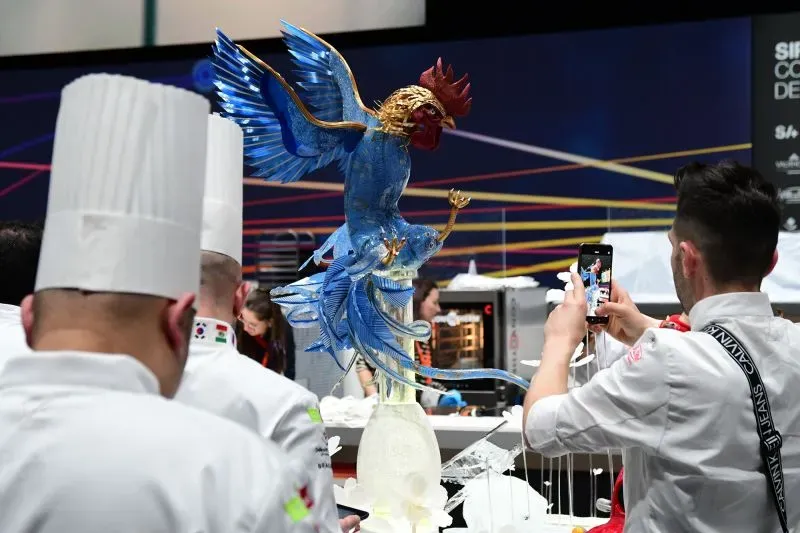 تمثال ملون لطائر زينة مصنوع من السكر، مع طهاة يرتدون زيًا رسميًا يتابعون الحدث في مسابقة كأس العالم للحلويات.