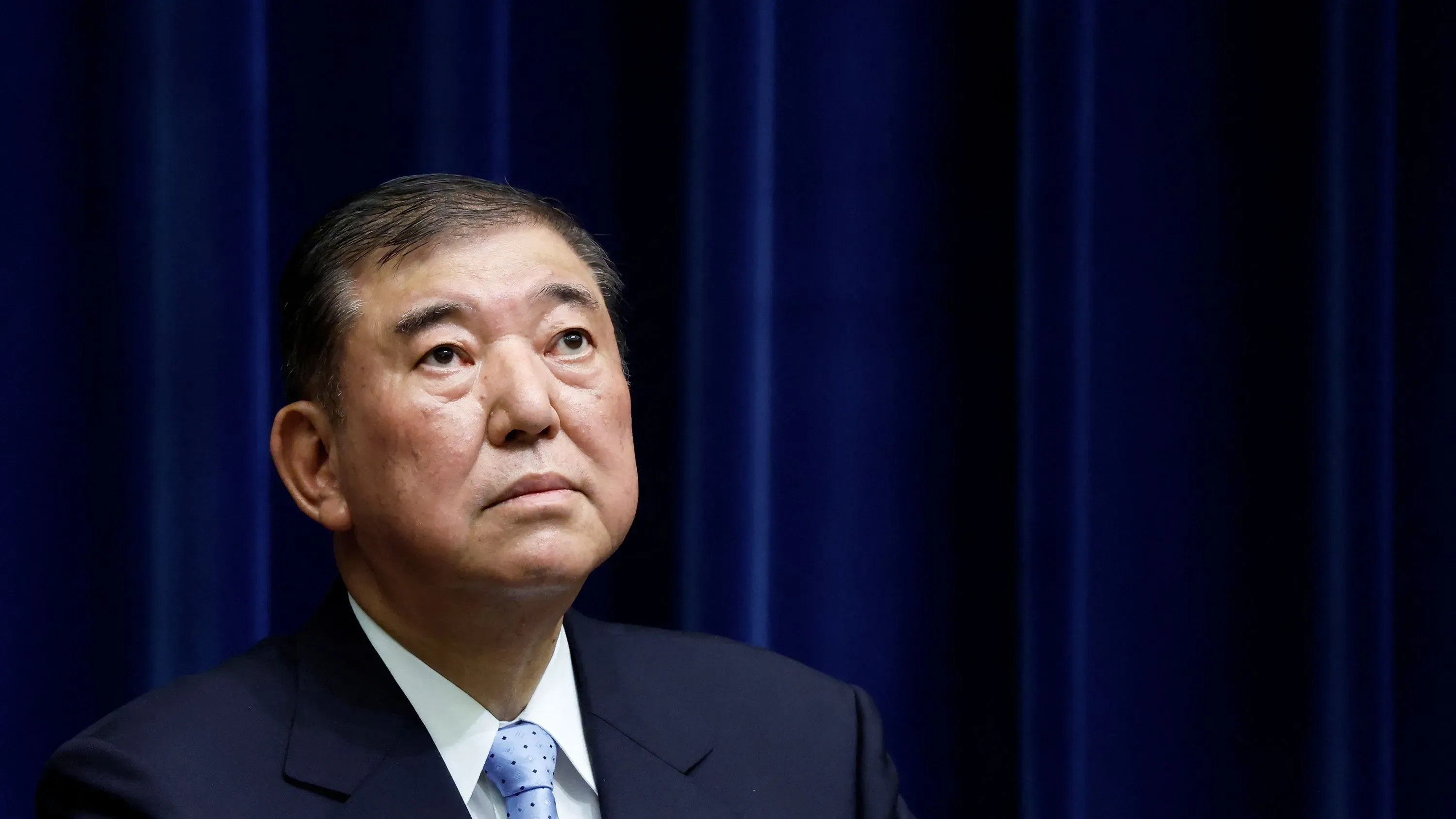 رجل في بدلة رسمية ينظر إلى الأعلى بقلق، مع خلفية زرقاء، في سياق سياسي متوتر في اليابان بعد الانتخابات.