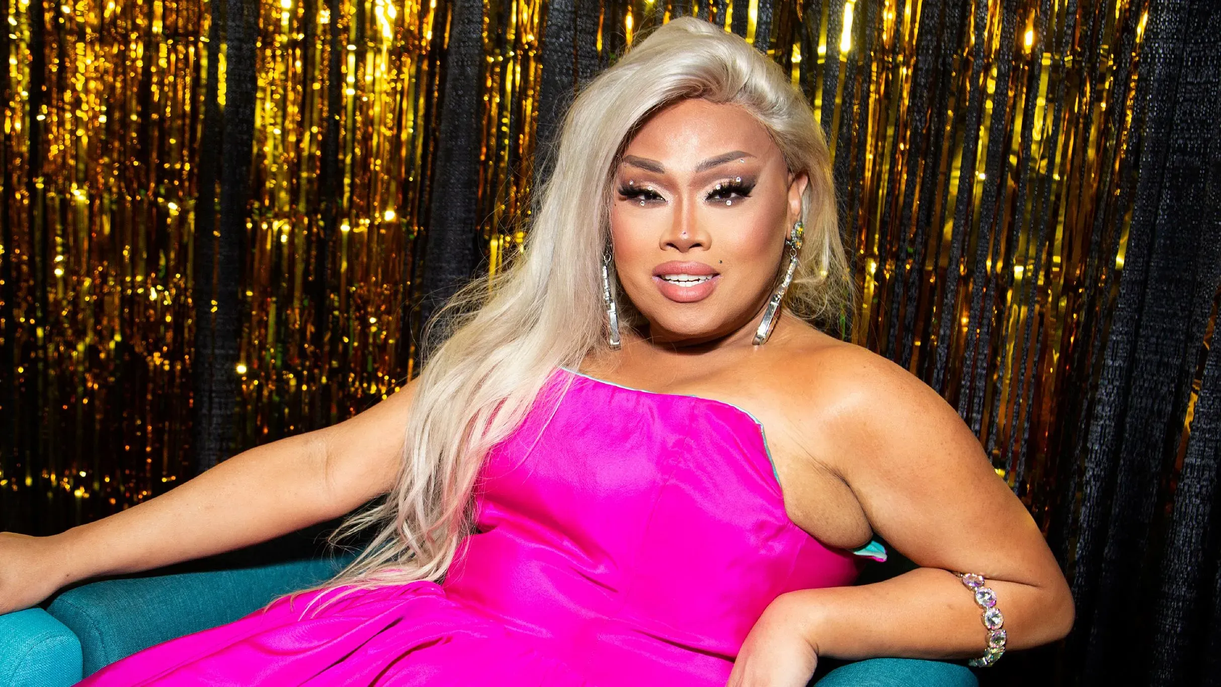 تظهر جيغلي كالينتي، فنانة وممثلة معروفة من برنامج "RuPaul's Drag Race"، ترتدي فستانًا ورديًا وتبتسم، محاطة بخلفية لامعة.