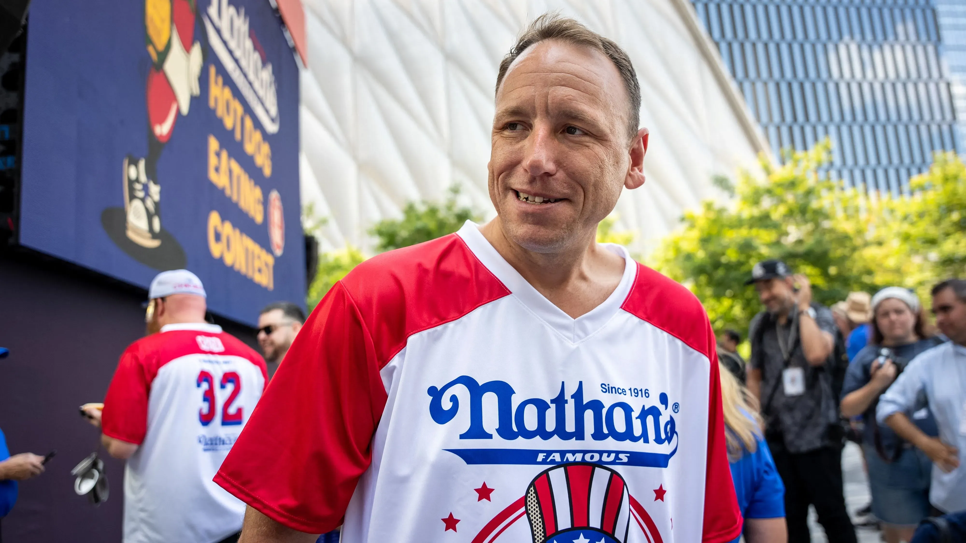 جوي تشيستنت مبتسم في مسابقة ناثانز لأكل الهوت دوغ، يرتدي قميصًا يحمل شعار المطعم، وسط حشد من المشجعين.