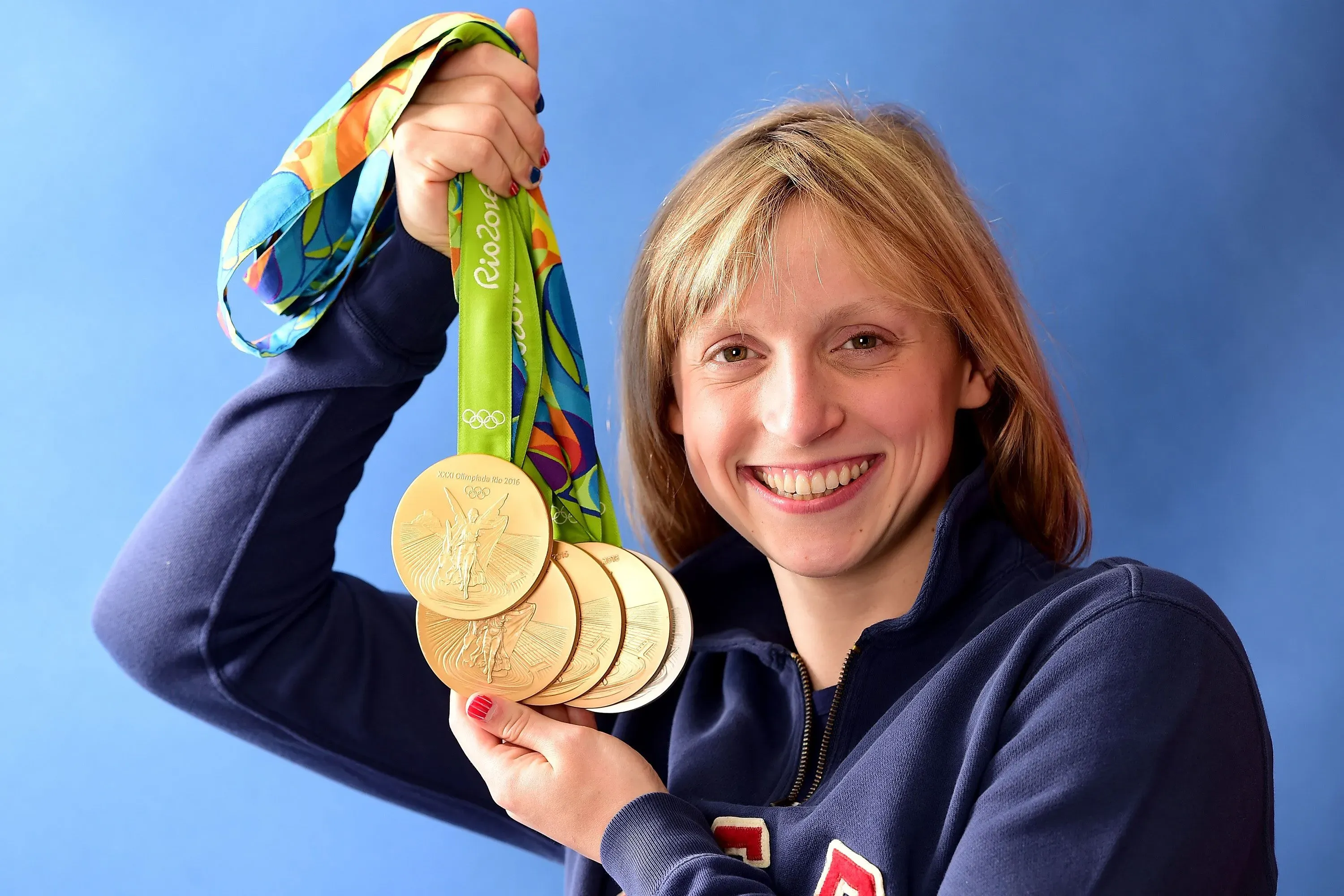 كاتي ليديكي تحمل خمس ميداليات ذهبية أولمبية مبتسمة، مع خلفية زرقاء، تعكس إنجازاتها في السباحة والتنافس في بطولة العالم.