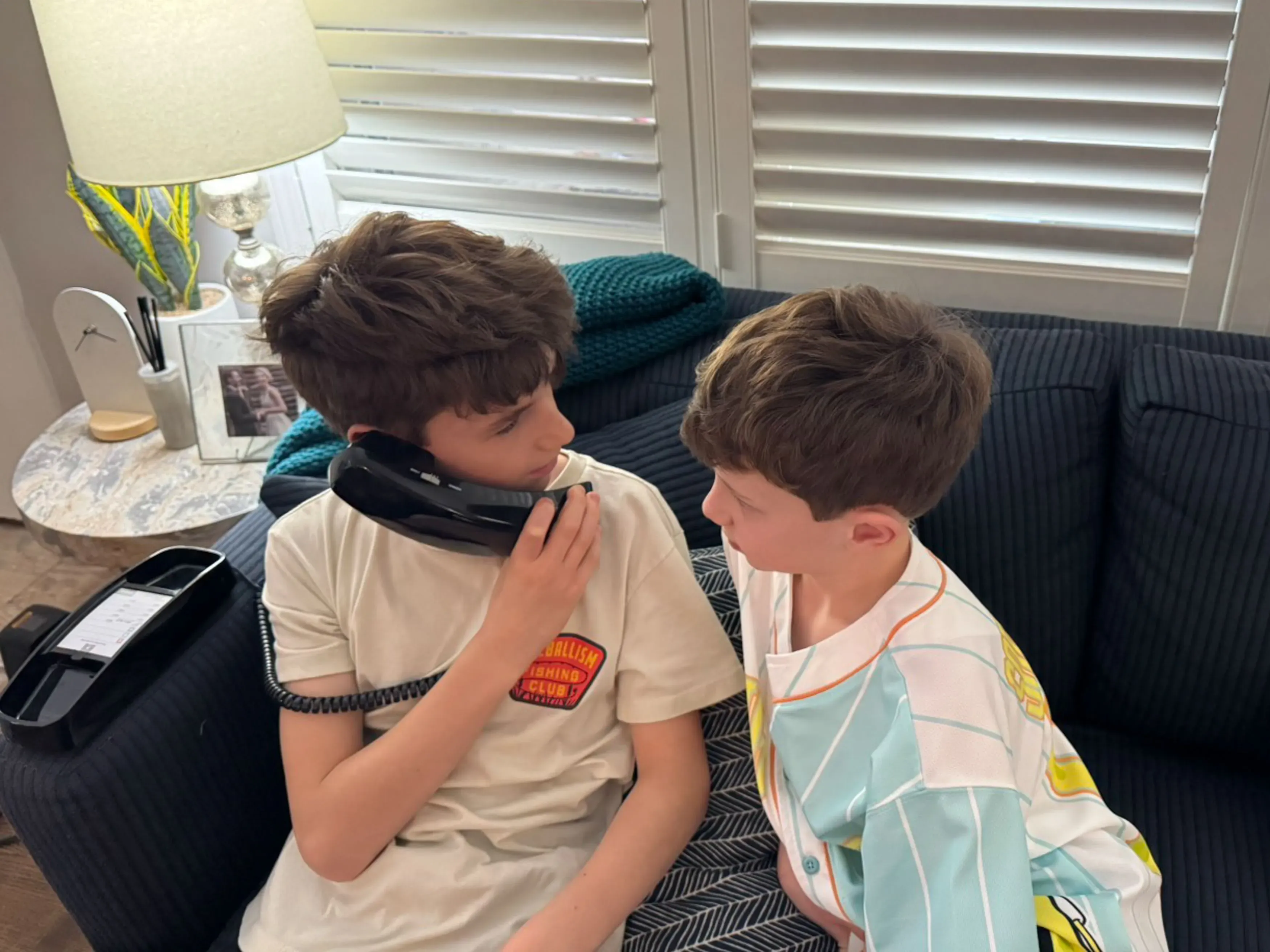 طفلان يجلسان على أريكة، يتحدثان عبر هاتف أرضي، مما يعكس أهمية التواصل التقليدي في عصر التكنولوجيا الحديثة.