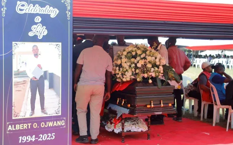 تجمع حشد من الناس حول نعش ألبرت أوجوانج، مع صورة له على لافتة تحت عنوان "الاحتفال بحياة". تظهر الأزهار والديكور في موقع الجنازة.