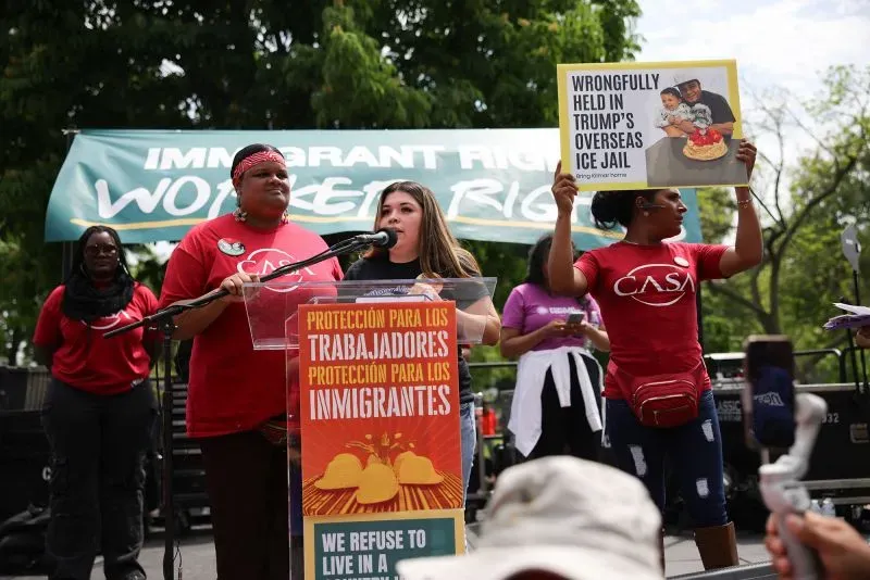 نساء يتحدثن في مظاهرة لدعم حقوق المهاجرين، مع لافتة تشير إلى احتجاز غير قانوني لشخص في سجن تابع لترامب.