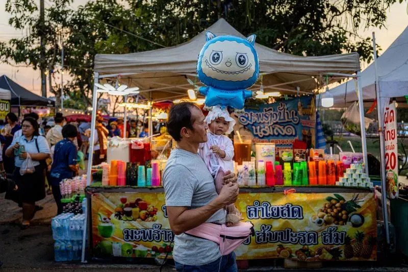 أب يحمل طفلة صغيرة ويحتفظ ببالون على شكل شخصية كرتونية في مهرجان، مع أكشاك ملونة في الخلفية.