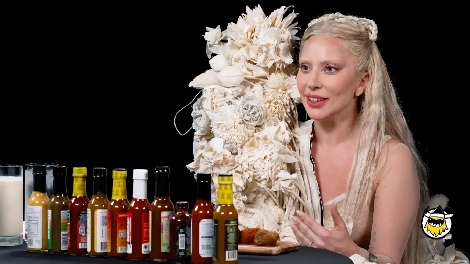 ليدي غاغا تتحدث في برنامج "Hot Ones" بينما تواجه أجنحة حارة، مرتدية فستاناً مميزاً، مع مجموعة من صلصات الفلفل أمامها.