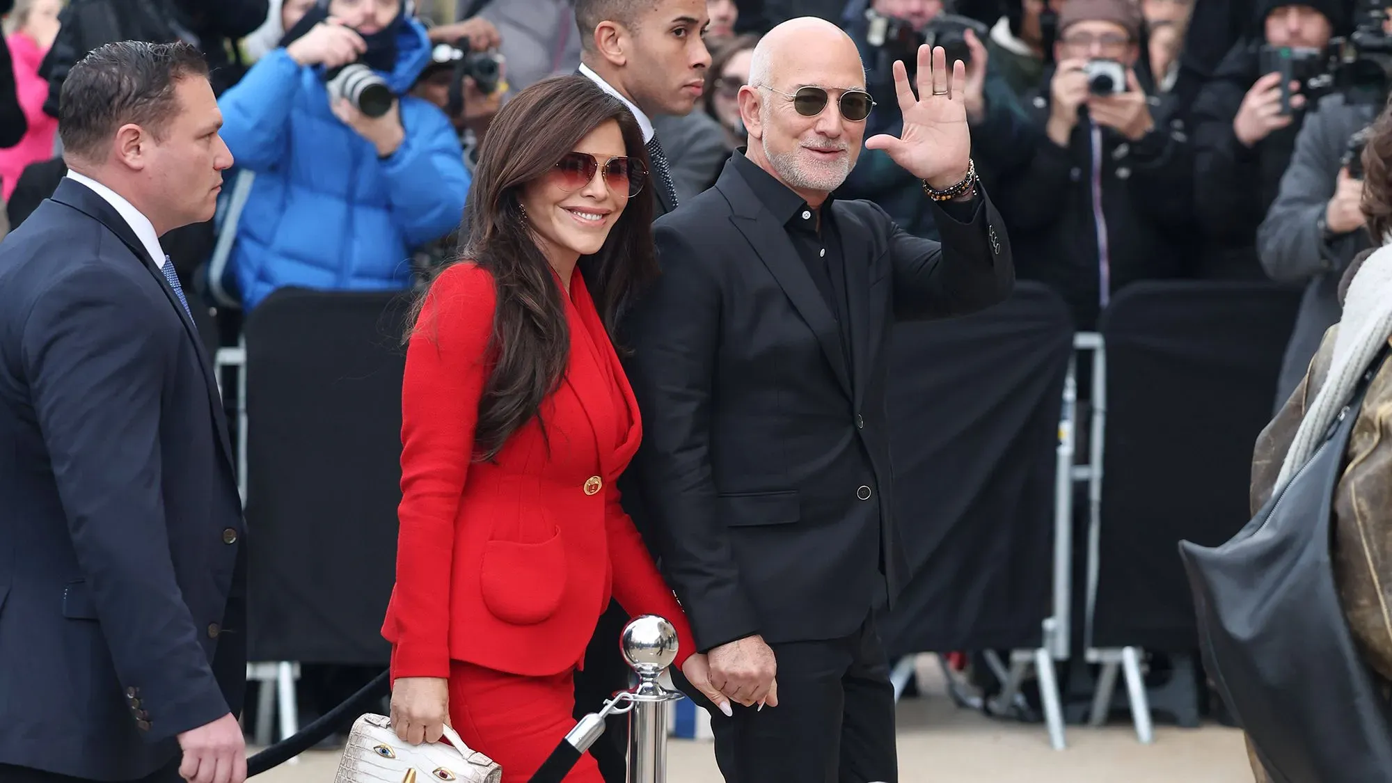 يظهر جيف بيزوس ولورين سانشيز بيزوس في عرض أزياء، حيث ترتدي سانشيز بدلة حمراء، بينما بيزوس يرتدي ملابس سوداء ونظارات شمسية.