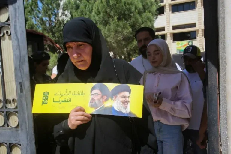امرأة ترتدي الحجاب تحمل لافتة دعماً لحزب الله، بينما يظهر عدد من الأشخاص خلفها في منطقة مزدحمة.