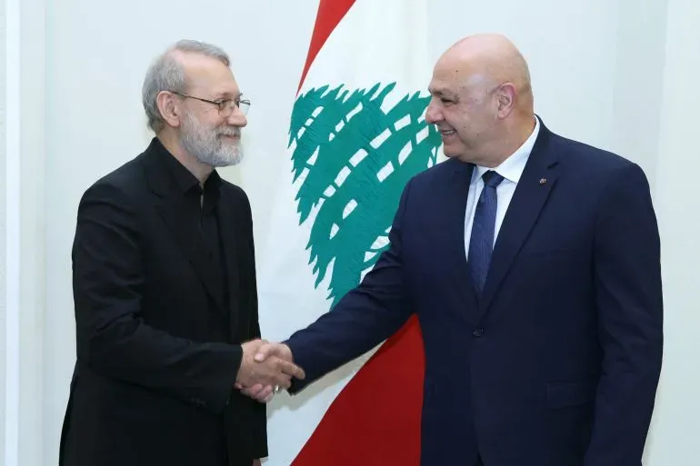 لقاء بين علي لاريجاني، رئيس المجلس الأعلى للأمن القومي الإيراني، ورئيس الوزراء اللبناني نواف سلام، مع العلم اللبناني خلفهما، حيث يتبادلان المصافحة.