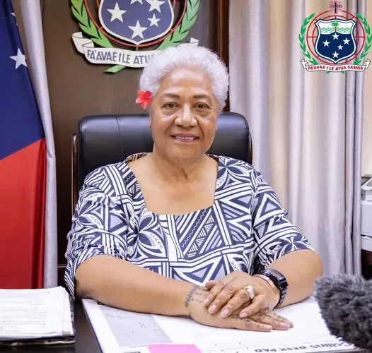 صورة لرئيسة وزراء ساموا، فيام نعومي مطاعفة، وهي تجلس في مكتبها، مرتدية زي تقليدي، مع العلم الوطني خلفها.