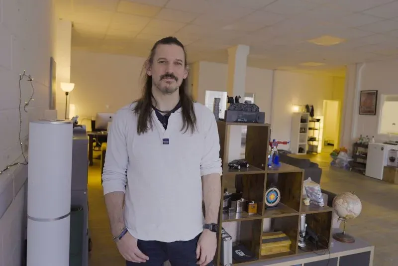 رجل يقف في مساحة مفتوحة لمكتب قديم في شرق لندن، حيث يظهر أثاث بسيط وأضواء دافئة، مع وجود مكونات تكنولوجية حوله.