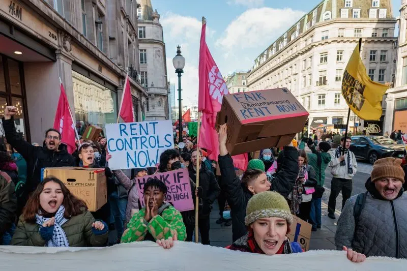 مظاهرة في لندن ضد ارتفاع الإيجارات، حيث يحمل المشاركون لافتات تدعو إلى تطبيق ضوابط للإيجار، مع الأعلام الوردية والخلفية الحضرية.
