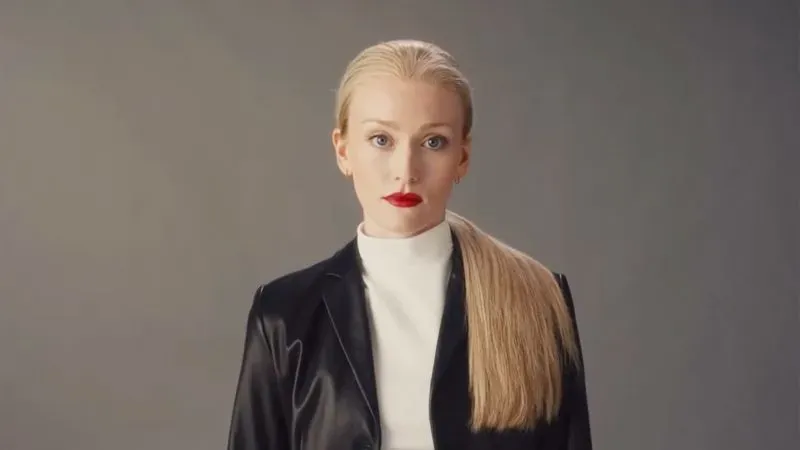 امرأة ذات شعر أشقر طويل ومربوط، ترتدي سترة جلدية سوداء وقميص أبيض، مع أحمر شفاه أحمر، تعبر عن الثقة.