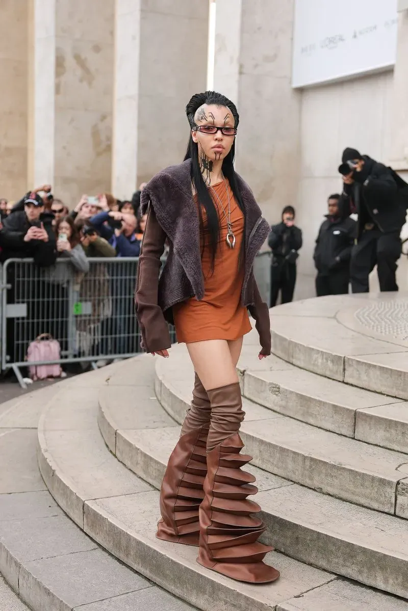 المغنية FKA Twigs تتألق في عرض ريك أوينز بباريس، مرتدية فستان قصير بني وحذاء كعب عالي فريد مع تفاصيل صناعية ومستقبلية.